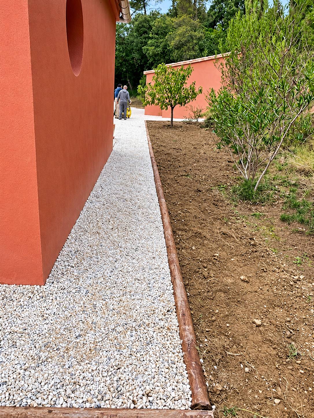 Une allée de gravier blanc bordée d'une bordure en bois longe un mur orangé, avec un jeune arbre planté dans la terre fraîchement remuée et des ouvriers au fond.