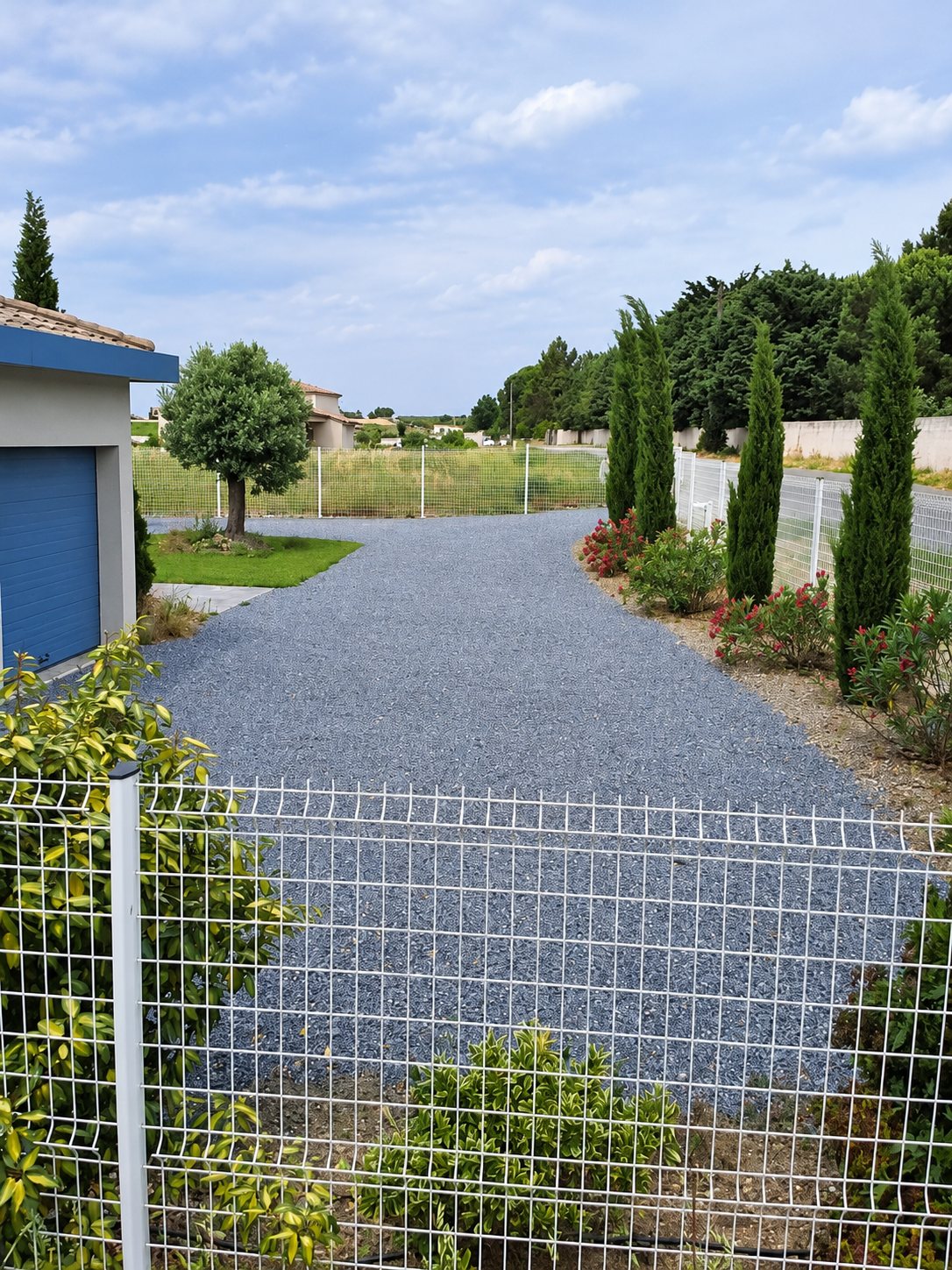 Une allée en gravier gris mène à un garage à porte bleue, bordée de cyprès, de lauriers-roses et d'une clôture en grillage blanc.