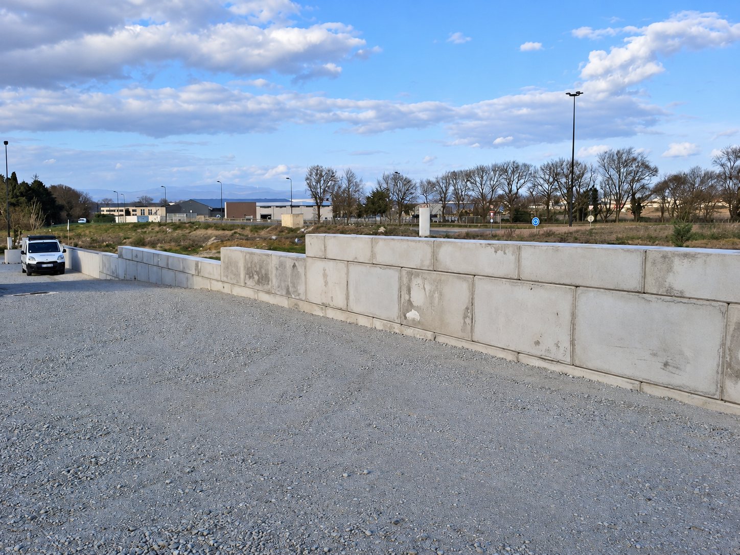 Un mur en gros blocs de béton gris borde une plateforme de gravier, avec une camionnette blanche et une zone d'activités en arrière-plan.