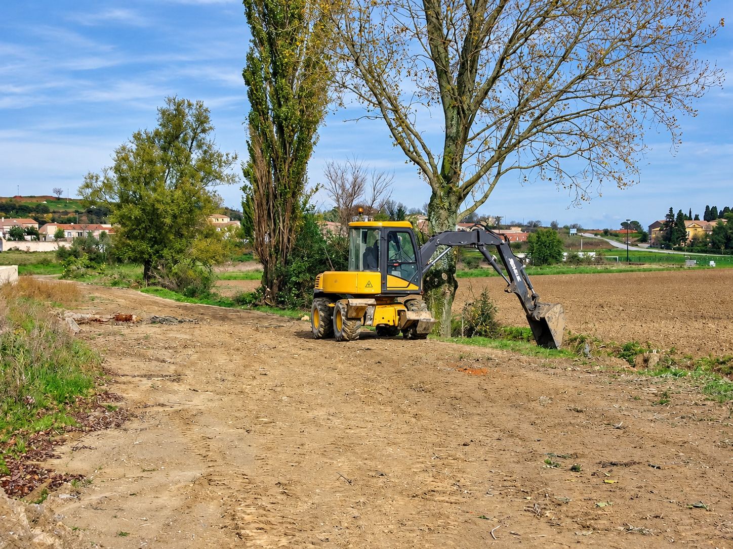 Une pelle mécanique jaune sur pneus travaille sur un chemin de terre au bord d'un champ, avec de grands arbres et un village en arrière-plan.