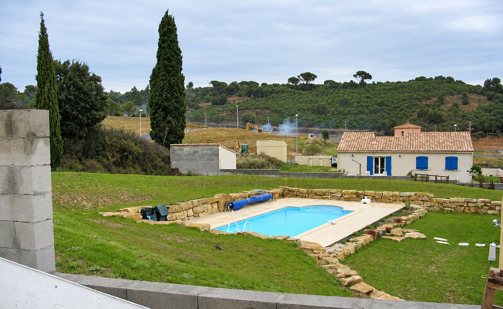 Une piscine rectangulaire avec margelles beige et murets de pierres sèches, aménagée sur une pelouse devant une maison à volets bleus.